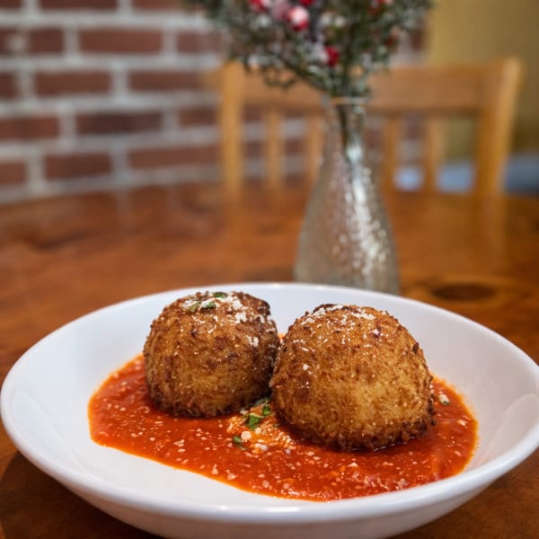 Two golden-brown arancini balls in a white bowl with red marinara sauce, sprinkled with cheese.