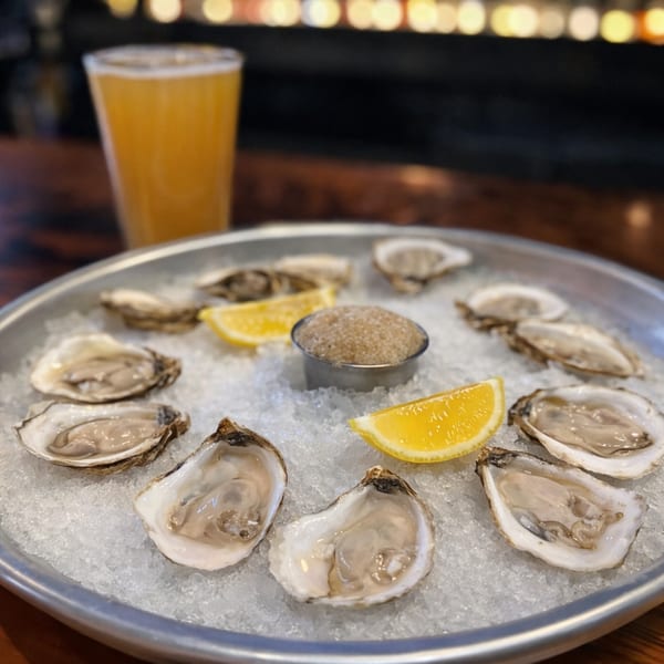 A dozen fresh oysters on a bed of ice with lemon wedges and mignonette, alongside a beer.